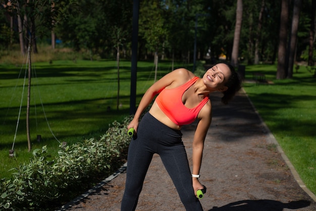 Mulher jovem e desportiva segurando halteres pequenos e se alongando após se exercitar em uma corrida ao ar livre