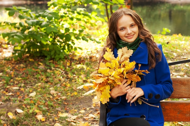 Mulher jovem e elegante com folhas de outono sentada no banco