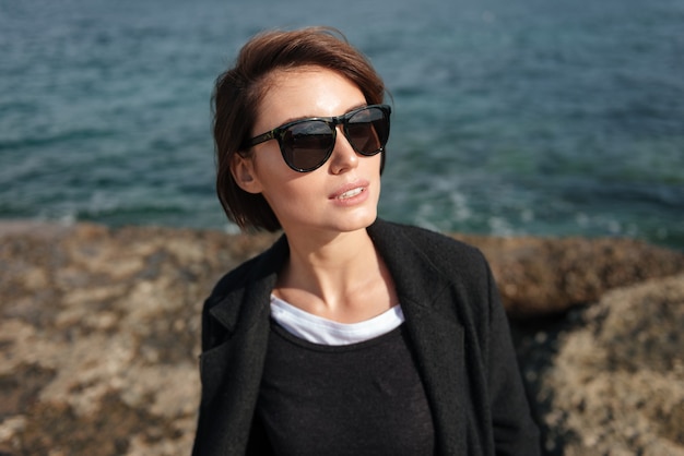 Mulher jovem e elegante com óculos de sol em pé perto do mar