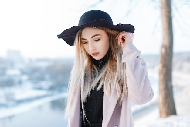 Mulher jovem e elegante com um chapéu vintage preto em estilo retro, com um casaco rosa moderno e um vestido de tricô, está na floresta em um dia quente de inverno