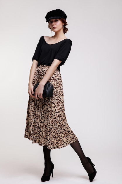 mulher jovem e elegante em saia longa de leopardo, top preto, chapéu, bolsa em fundo branco