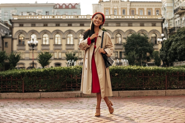 Mulher jovem e elegante morena com um casaco impermeável bege, vestido midi vermelho e boina, sorrindo e fazendo poses de bom humor lá fora