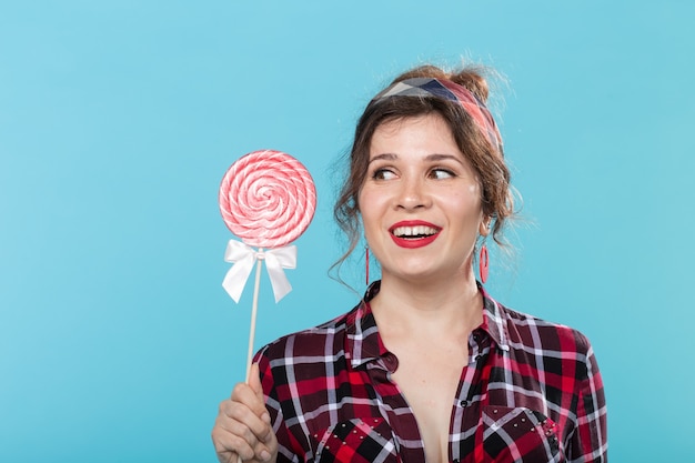 Mulher jovem e encantadora com roupas retrô, segurando pirulitos coloridos nas mãos e lambendo um deles posando sobre fundo azul. Conceito de amor por doces e festas. Copyspace