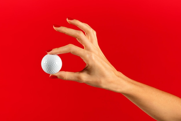 Foto mulher jovem e esbelta anônima segurando uma bola de golfe com unhas polidas, índice esbelto e dedos do polegar enquanto demonstra contra fundo vermelho na luz