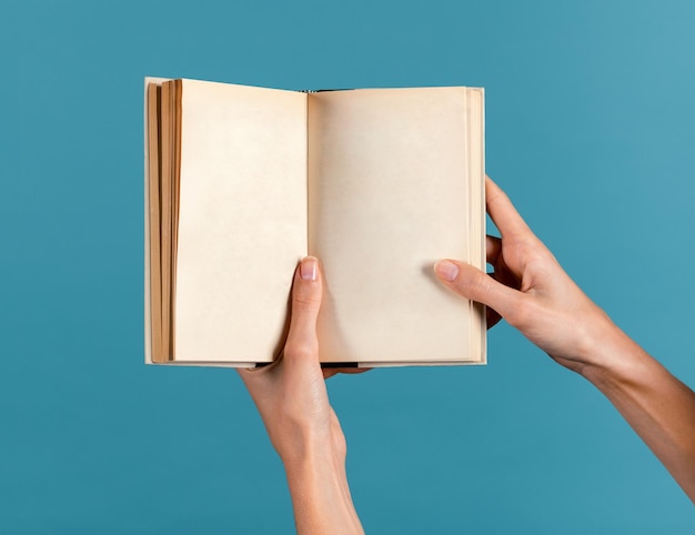 Mulher jovem e esguia anônima com dedos manicuredos segurando páginas vazias livro encadernado em capa dura à luz e demonstrando contra fundo azul