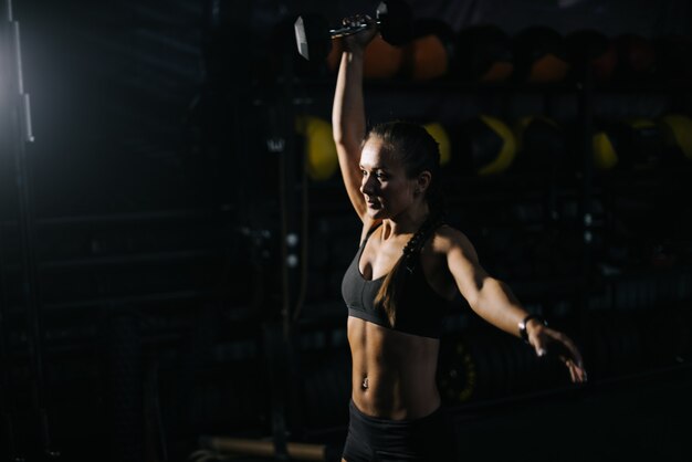 Mulher jovem e esportiva com corpo atlético perfeito, vestindo uma roupa esportiva preta levantando o kettlebell acima da cabeça