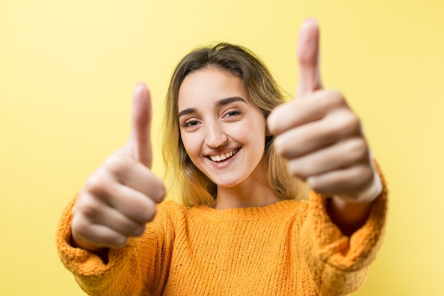 Mulher jovem e feliz, branca, com um suéter laranja, fazendo sinal de polegar para cima e sorrindo. Bom trabalho