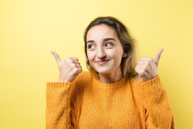 Mulher jovem e feliz, branca, com um suéter laranja, fazendo sinal de polegar para cima e sorrindo. bom trabalho