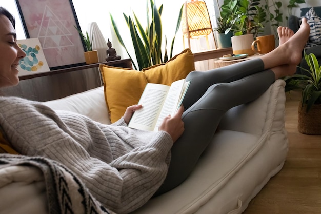 Mulher jovem e feliz caucasiana lendo um livro em casa. Relaxando no sofá. Conceito de estilo de vida.