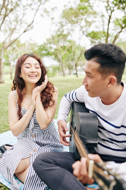 Mulher jovem e feliz e animada aplaudindo o namorado cantando e tocando violão quando eles estão fazendo um piquenique romântico no parque