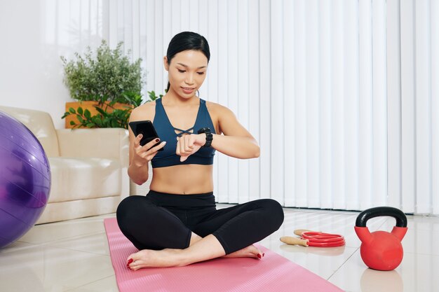 Mulher jovem e feliz em forma verificando aplicativos em smartwatch e smartphone antes de se preparar para fazer exercícios em casa