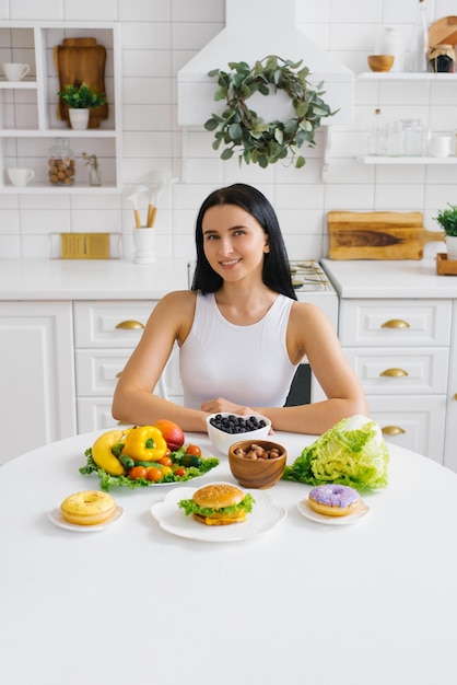 Mulher jovem e feliz está comendo comida saudável enquanto está sentado em uma mesa com ingredientes verdes frescos dentro de casa