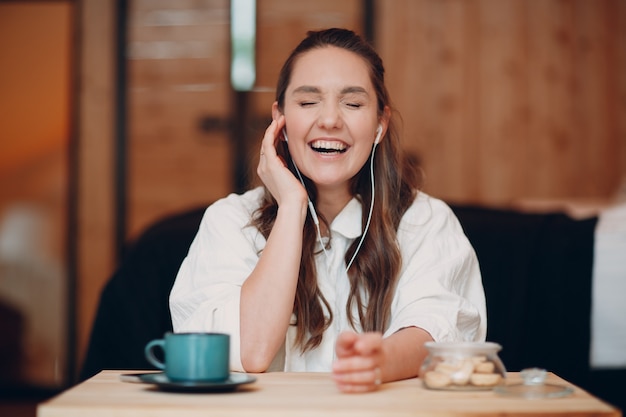 Mulher jovem e feliz rindo sentada à mesa em casa atrás do laptop do computador e falando na videochamada garota feminina com fones de ouvido móveis portáteis falando online pela webcam dentro de casa