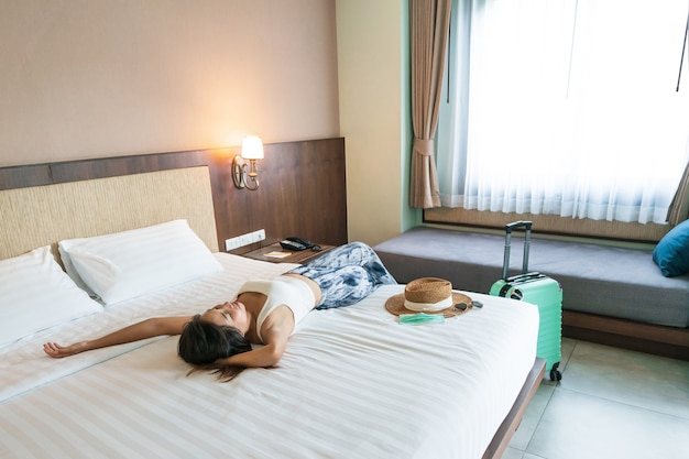 Mulher jovem e feliz viajante asiática relaxando na cama com chapéu e máscara médica na cama em um quarto de hotel