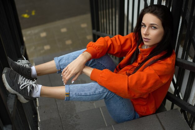 Mulher jovem e hippie na moda sentada em uma escada de verão com uma jaqueta vermelha e calça jeans azul