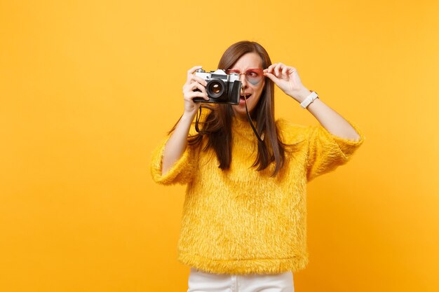 Mulher jovem e intrigada chocada segurando óculos de coração e tirando fotos na câmera fotográfica vintage retrô isolada em fundo amarelo brilhante. emoções sinceras de pessoas, conceito de estilo de vida. área de publicidade.