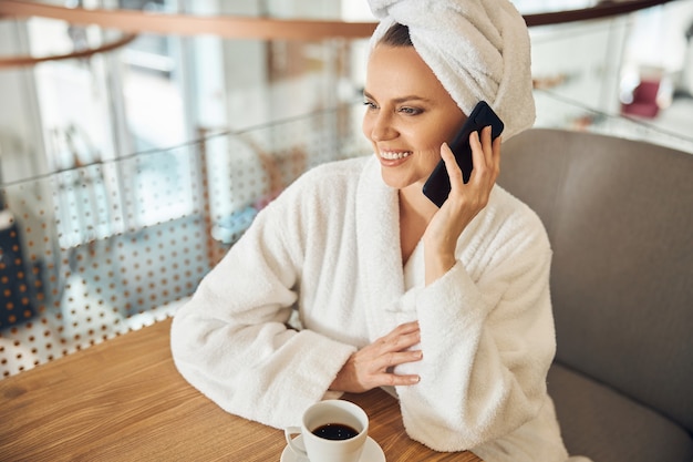 Mulher jovem e moderna sorridente, apoiando os cotovelos em uma mesa de madeira durante a conversa por telefone