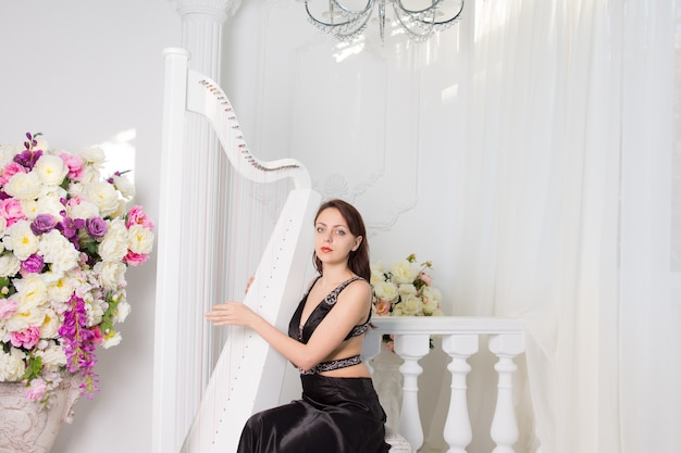 Mulher jovem e muito elegante tocando harpa em um concerto clássico ou recital sentada em um palco branco decorado com flores frescas coloridas