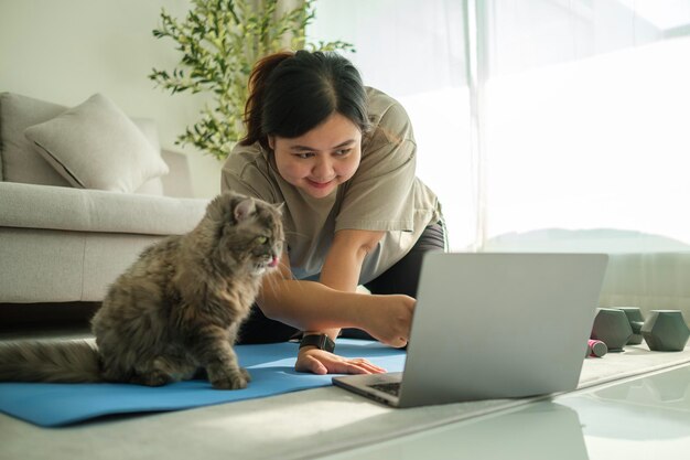 Mulher jovem e obesa sorridente a exercitar-se na sala de estar com um gato bonito a ver online