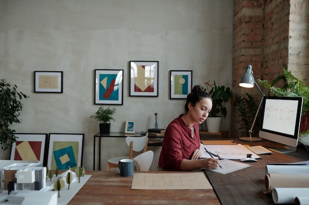 Mulher jovem e ocupada de raça mista com coque de cabelo sentada à mesa e desenhando um esboço enquanto trabalhava no escritório com fotos na parede