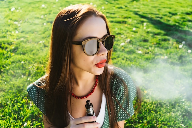 Mulher jovem e positiva de óculos escuros sentada no gramado com grama verde brilhante, fumando, pensativa