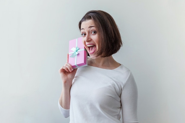 Mulher jovem e positiva segurando uma pequena caixa de presente rosa isolada no branco, parecendo feliz e animada