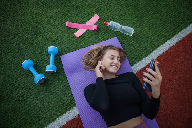 Mulher jovem e saudável está descansando deitada sobre o tapete e ouvir música. Treinamento ao ar livre. Fitness, conceito de esporte. Estilo de vida saudável
