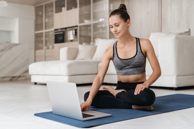 Mulher jovem e sorridente aprendendo exercícios de ioga sentada na esteira em posição de lótus assistindo a um vídeo no laptop