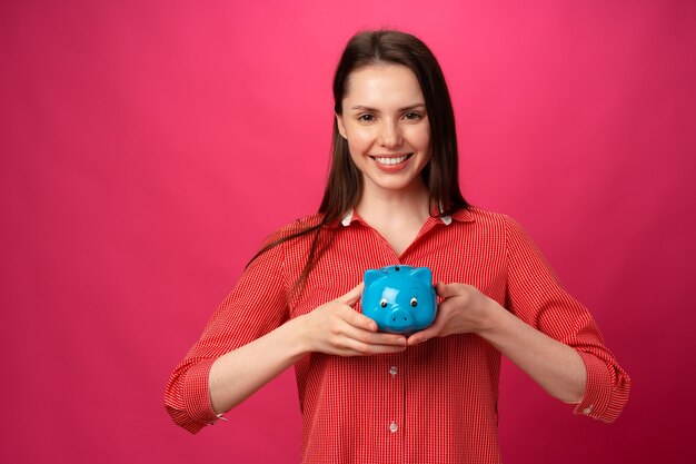 Mulher jovem e sorridente feliz segurando o cofrinho azul contra um fundo rosa