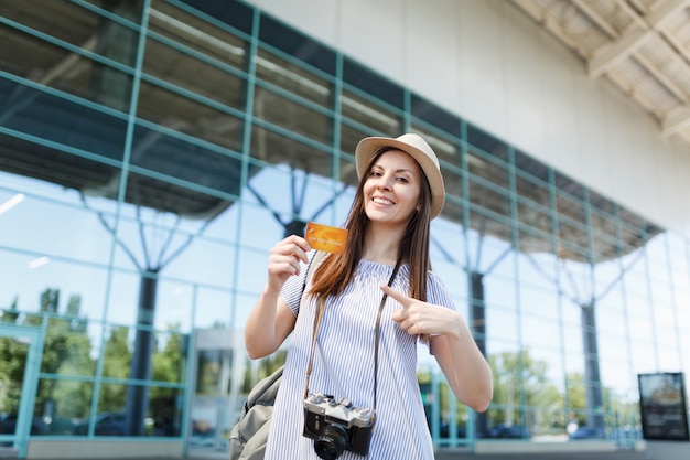 Mulher jovem e sorridente viajante turista com câmera fotográfica vintage retrô, apontando o dedo indicador no cartão de crédito no aeroporto internacional