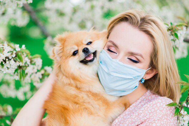 Mulher jovem e spitz vermelho com uma máscara médica no rosto na natureza em um dia de primavera. Pandemia de coronavírus, quarentena.