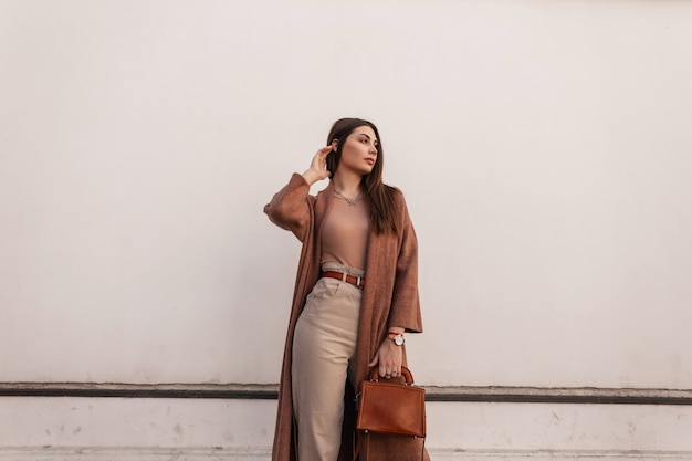 Foto mulher jovem elegante bonita em elegante casaco em calças com bolsa de couro marrom elegante posando perto de prédio branco vintage na rua. garota atraente urbana descansa ao ar livre. roupa casual de primavera na moda