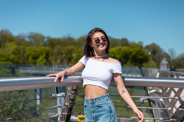 Mulher jovem elegante com blusa casual branca e calça jeans posando na ponte da cidade em um dia ensolarado e quente