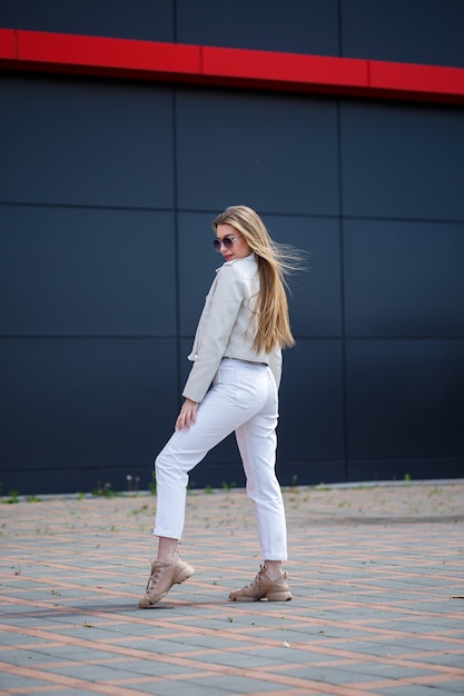 Mulher jovem elegante com longos cabelos loiros de aparência europeia com um sorriso no rosto. Menina com uma jaqueta branca e calça jeans branca um dia ensolarado de verão quente no fundo de um edifício cinza