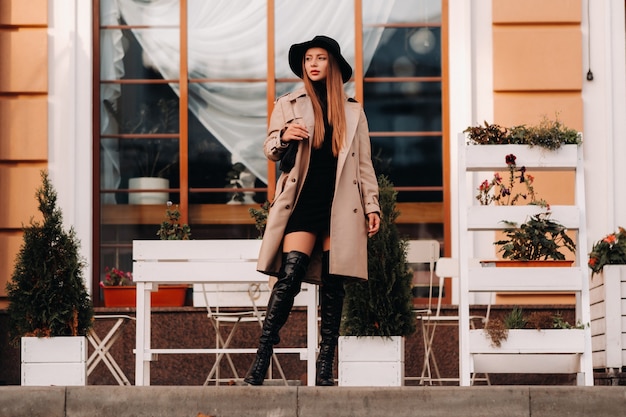 Mulher jovem elegante com um casaco bege com um chapéu preto em uma rua da cidade. Moda feminina de rua. Roupas de outono. Estilo urbano.