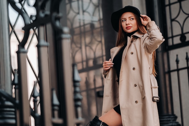 Mulher jovem elegante com um casaco bege com um chapéu preto em uma rua da cidade. Moda feminina de rua. Roupas de outono. Estilo urbano.