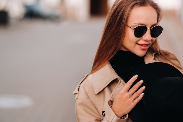 Mulher jovem elegante com um casaco bege e um chapéu preto nas mãos e óculos em uma rua da cidade. Moda feminina de rua. Roupas de outono. Estilo urbano.