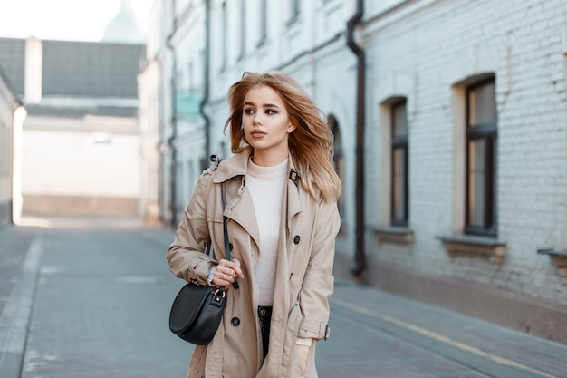 Mulher jovem elegante com um casaco leve de primavera em uma camiseta branca com uma bolsa de couro preto elegante andando pela rua no contexto de um antigo edifício de tijolos. Linda garota europeia elegante