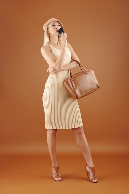 Mulher jovem elegante e pensativa posando com bolsa de couro e smartphone