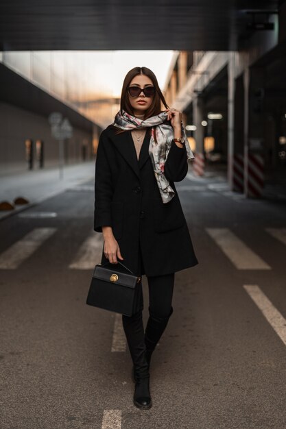 Mulher jovem elegante em óculos de sol com lenço de seda elegante na cabeça em casaco preto elegante em jeans com botas com bolsa fica no asfalto da cidade. modelo de garota de negócios caminha na estrada na rua.
