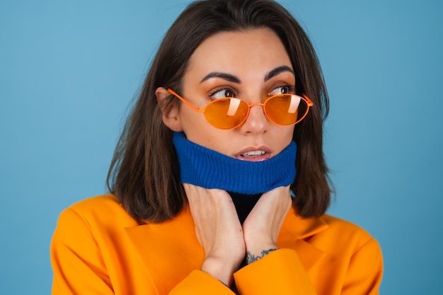 Mulher jovem elegante em uma jaqueta grande e comprida em uma parede azul com óculos laranja brilhante elegantes posando