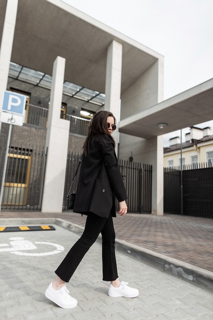 Mulher jovem elegante urbana no blazer da juventude vintage em óculos de sol da moda com bolsa de couro se passando perto de um edifício moderno na cidade. Hipster de garota na moda atraente em jaqueta preta de moda na rua.