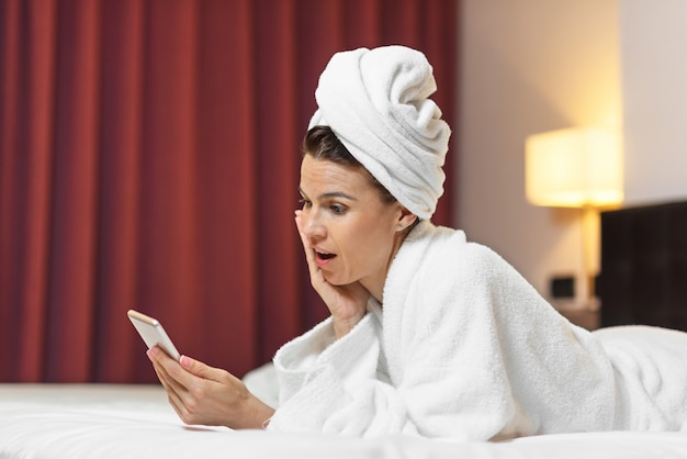 Mulher jovem, em, bathrobe, mentindo, em, quarto hotel, usando, telefone móvel, relaxado, após, fazendo exame um banho