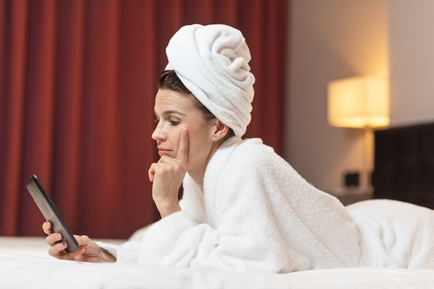 Mulher jovem, em, bathrobe, mentindo, em, quarto hotel, usando, telefone móvel, relaxado, após, fazendo exame um banho