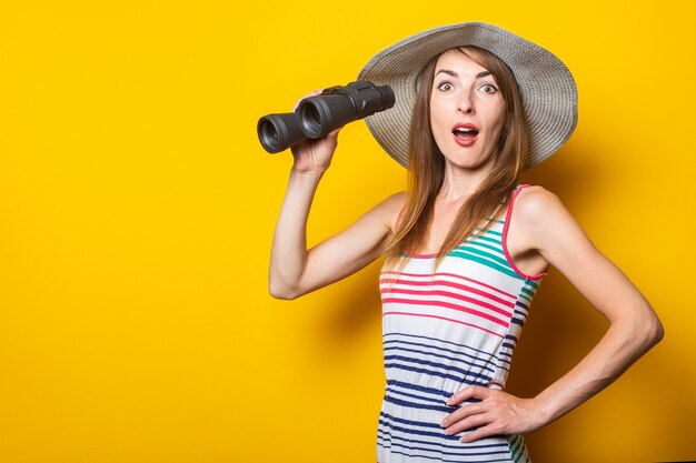 Mulher jovem em estado de choque, surpresa, com um chapéu e um vestido listrado segura um binóculo em um espaço amarelo.