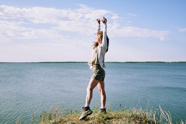 Mulher jovem em êxtase com os braços levantados e os olhos fechados enquanto está de pé na margem do rio, perto da água, desfrutando da solidão