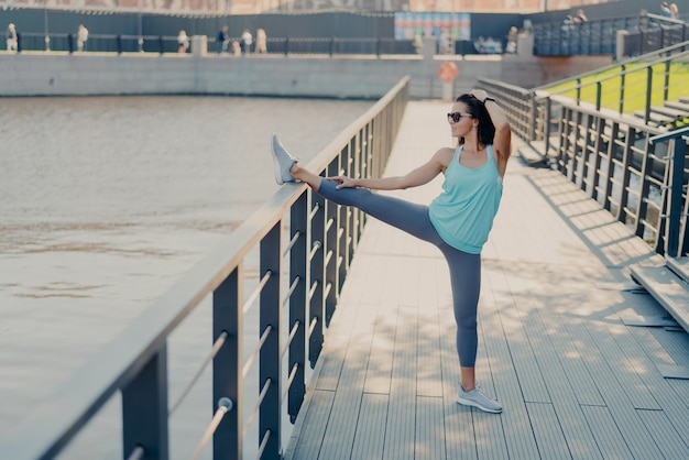 Mulher jovem em forma com cabelo escuro estica as pernas na cerca se aquece antes de correr usa óculos de sol, camiseta, legging e tênis se prepara para poses de treinamento cardiovascular ao ar livre estando em boa forma física