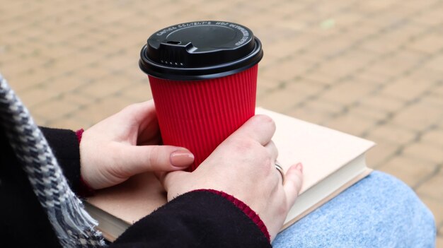 Mulher jovem em jeans, casaco e lenço, em um banco do parque. uma mulher está lendo um livro e tomando café ou outra bebida quente sozinha ao ar livre. fechar-se. o conceito de honra, estudo, lazer e recreação.