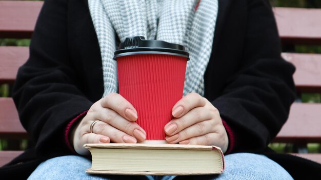 Mulher jovem em jeans, casaco e lenço, em um banco do parque. Uma mulher está lendo um livro e tomando café ou outra bebida quente sozinha ao ar livre. Fechar-se. O conceito de honra, estudo, lazer e recreação.