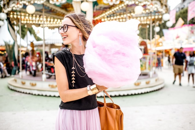 Mulher jovem em pé com algodão doce rosa ao ar livre em frente ao carrossel no parque de diversões